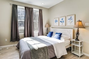 Ein Schlafzimmer mit einem Bett, zwei Nachttischlampen, einem Nachttisch, einem Fenster mit Vorhängen und zwei an der Wand befestigten Blumenbildern.