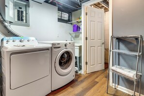 A laundry area with a front-loading washing machine, a door leading to a bathroom, and a shelving unit with cleaning supplies. - 8 Mi to Dtwn Detroit! Apt w/ Shared Yard (Dearborn)