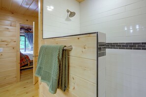 Interior em madeira com chuveiro, uma toalha pendurada na parede e um vislumbre de uma cama com um cobertor xadrez.