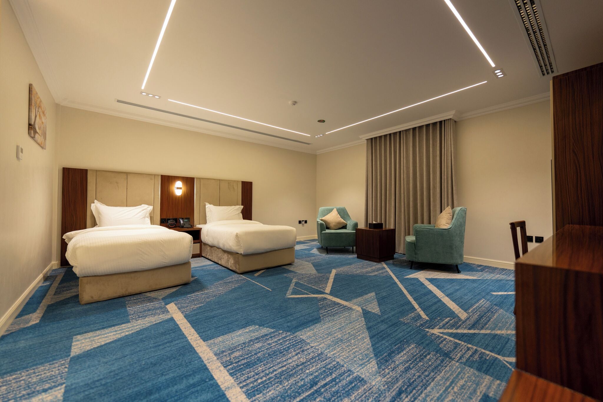 Habitación de hotel con dos camas, un escritorio, una silla, un ventanal grande con cortinas y una alfombra azul.