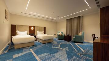 Habitación de hotel con dos camas, un escritorio, una silla, un ventanal grande con cortinas y una alfombra azul.