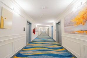 Un pasillo de hotel con alfombra estampada, puertas a ambos lados y un cuadro en la pared derecha.