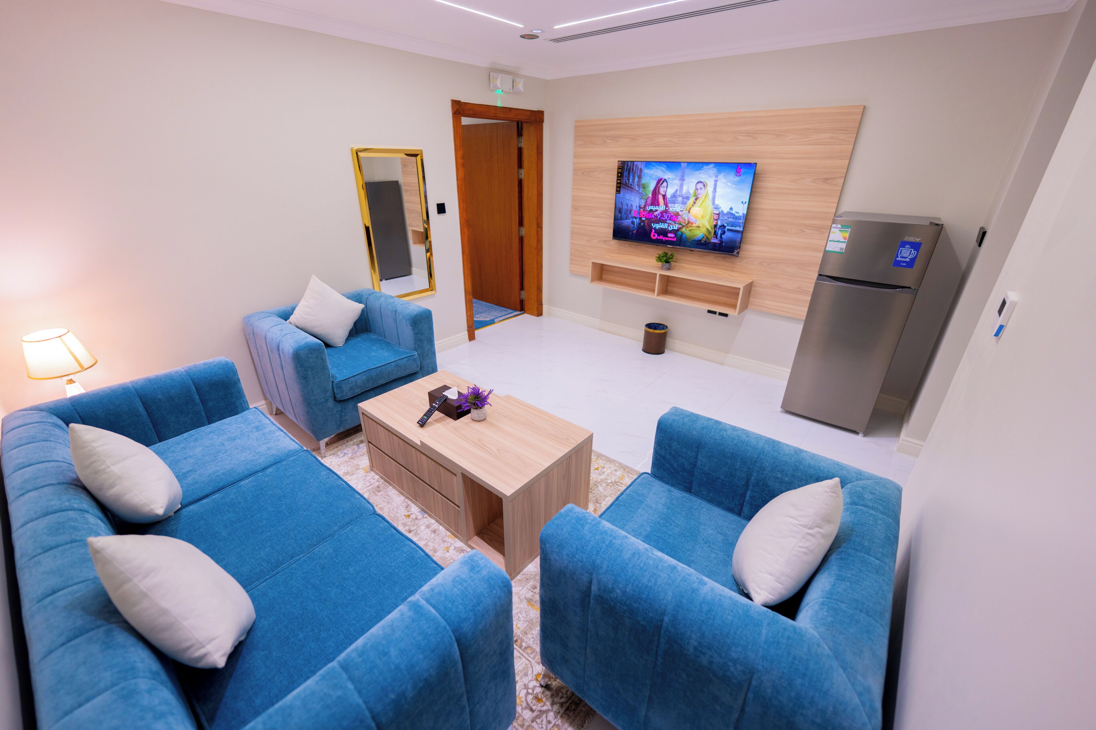 Un salon moderne avec un canapé bleu, une table basse en bois, un téléviseur à écran plat fixé au mur et un réfrigérateur.