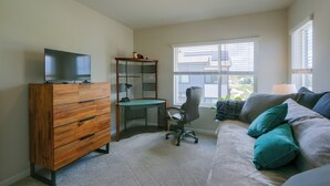 Une pièce comprenant une commode en bois, un bureau avec une chaise, un téléviseur posé sur la commode, une fenêtre avec des stores et un canapé avec des coussins.