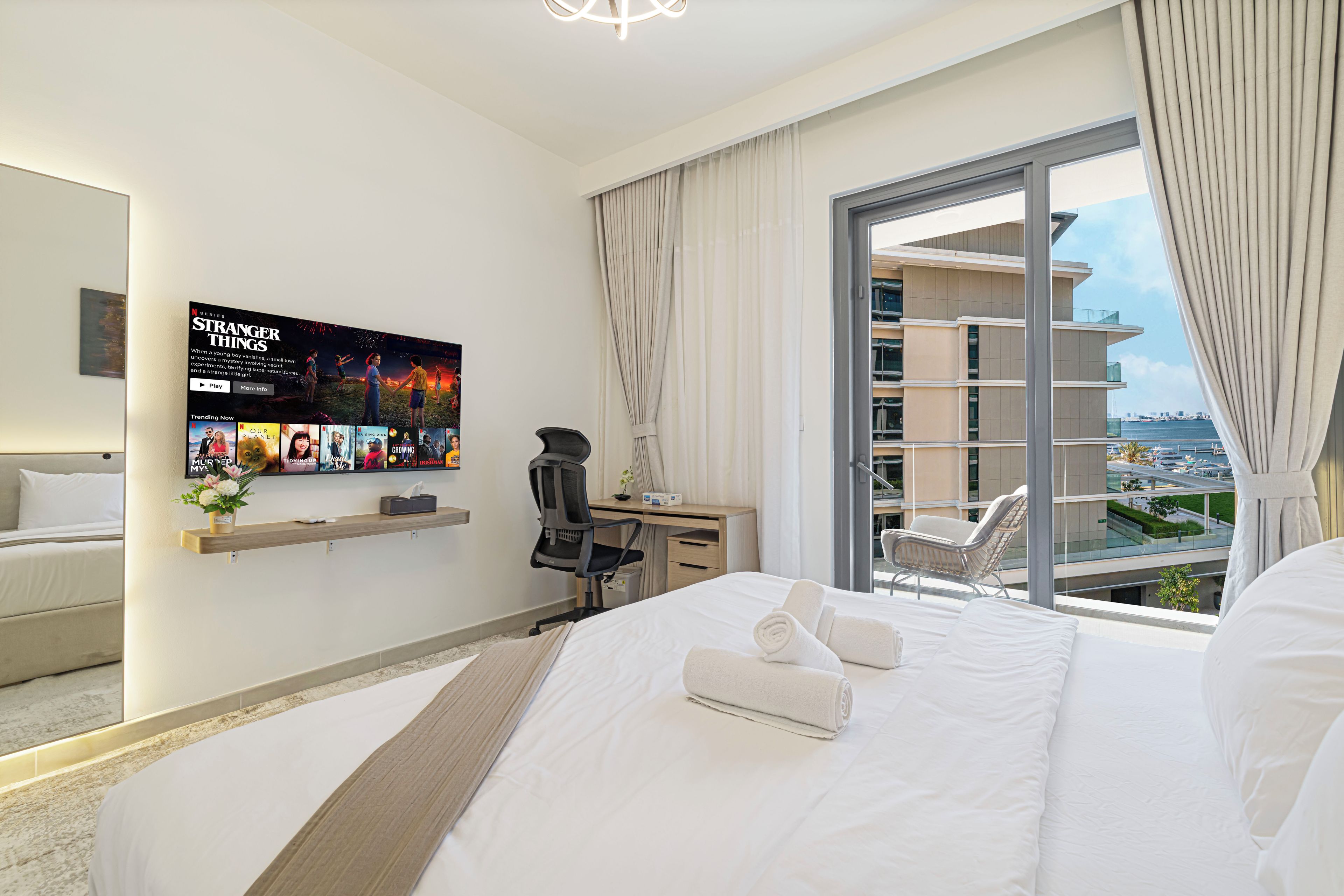 A modern hotel room with a large bed, a desk, a chair, a TV mounted on the wall, and a balcony with a view of the city.