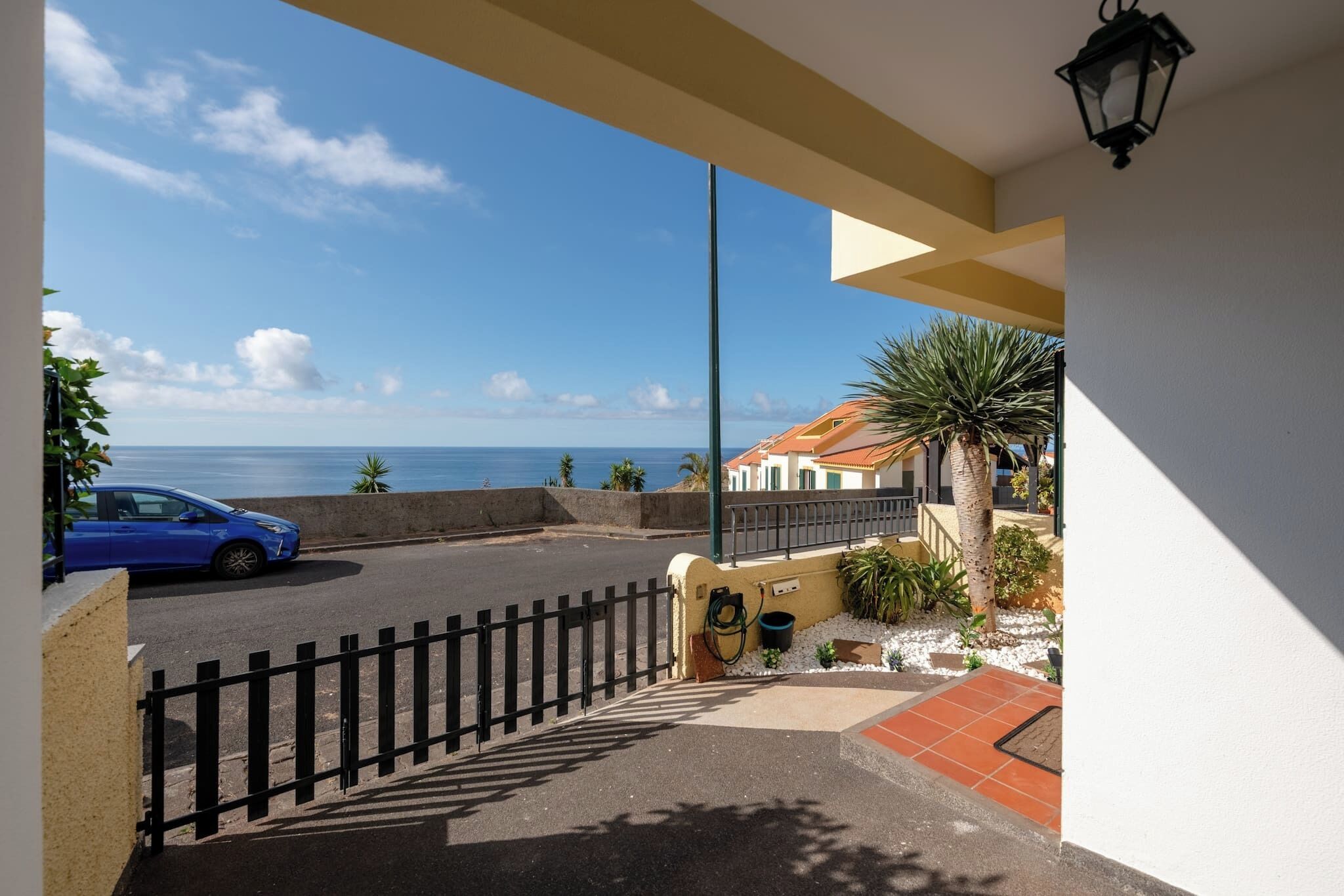 A balcony with a view of the sea, a parked car, and a building with a tiled roof.