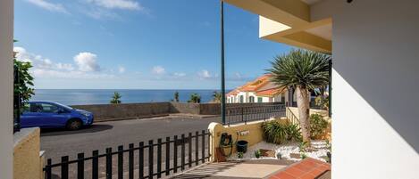 A balcony with a view of the sea, a parked car, and a building with a tiled roof.