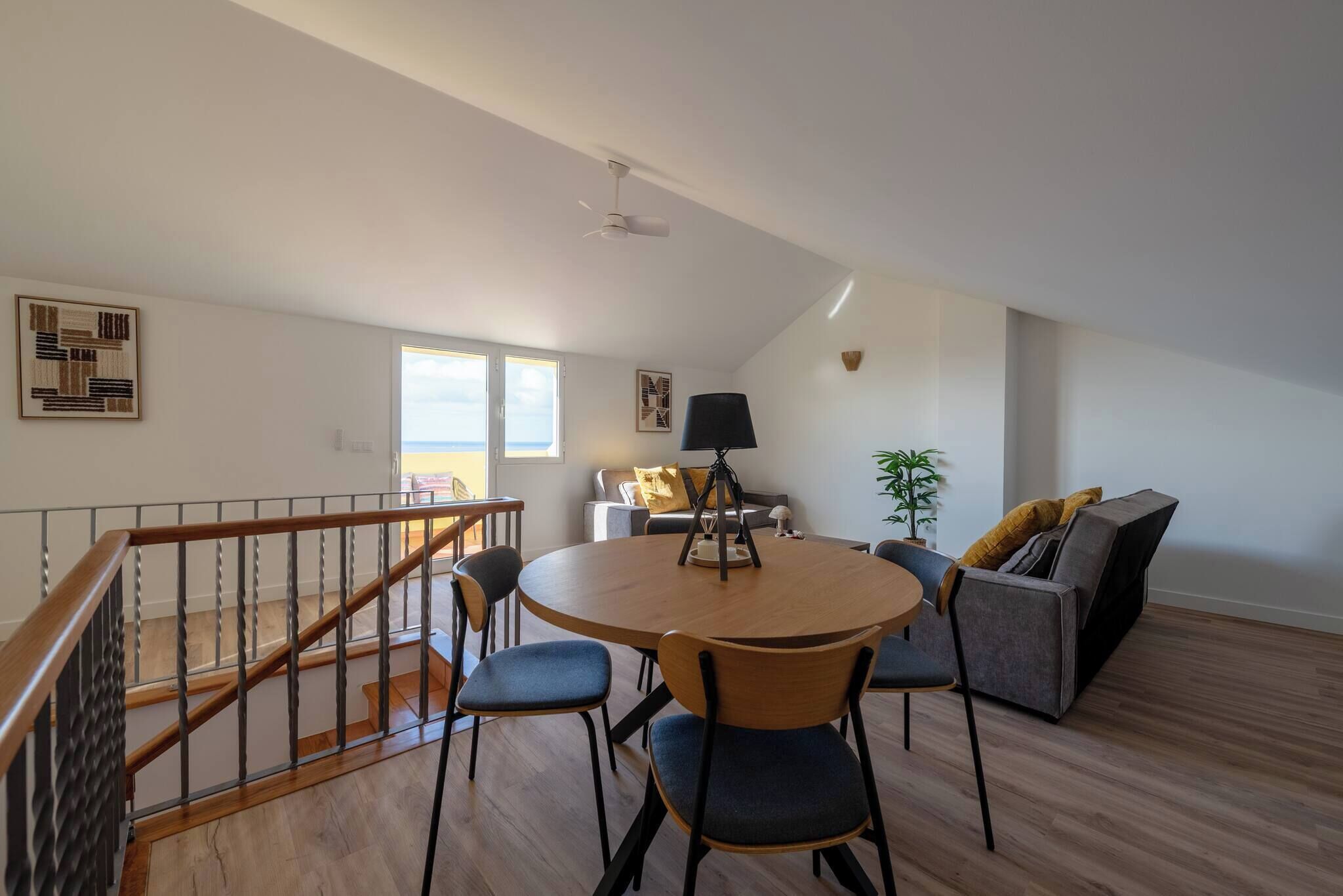 A spacious living area with a round wooden table, blue chairs, a grey sofa, and a staircase with a wooden railing.