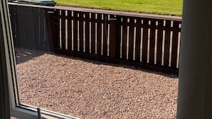 A view through a window of a gravel area with a wooden fence, a grassy field, and a body of water in the distance. - Loch linnhe view house, Fort William (Caol)