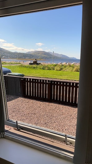 A view through a window of a gravel area with a wooden fence, a grassy field, and a body of water in the distance. - Loch linnhe view house, Fort William (Caol)