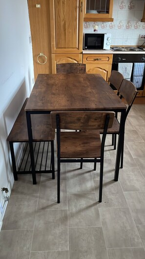 Uma área de refeições com mesa e cadeiras de madeira, uma cozinha com armários de madeira e micro-ondas, e piso revestido de azulejos.