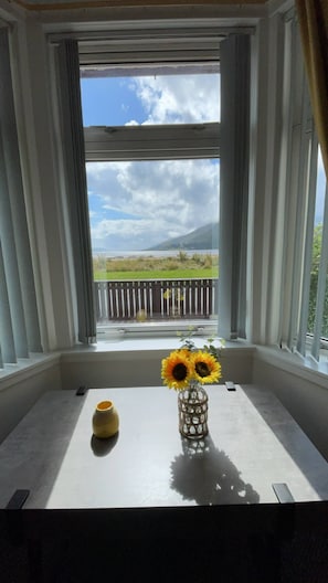 A room with a large window, a table with a vase of sunflowers, and a yellow object on the table. - Loch linnhe view house, Fort William (Caol)