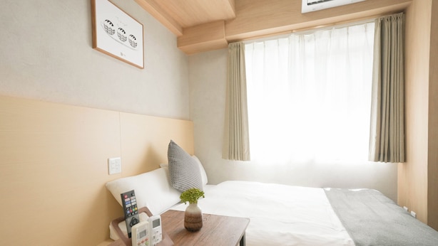 Una habitación de hotel con cama, mesita de noche con teléfono, florero con planta y ventana con cortinas.