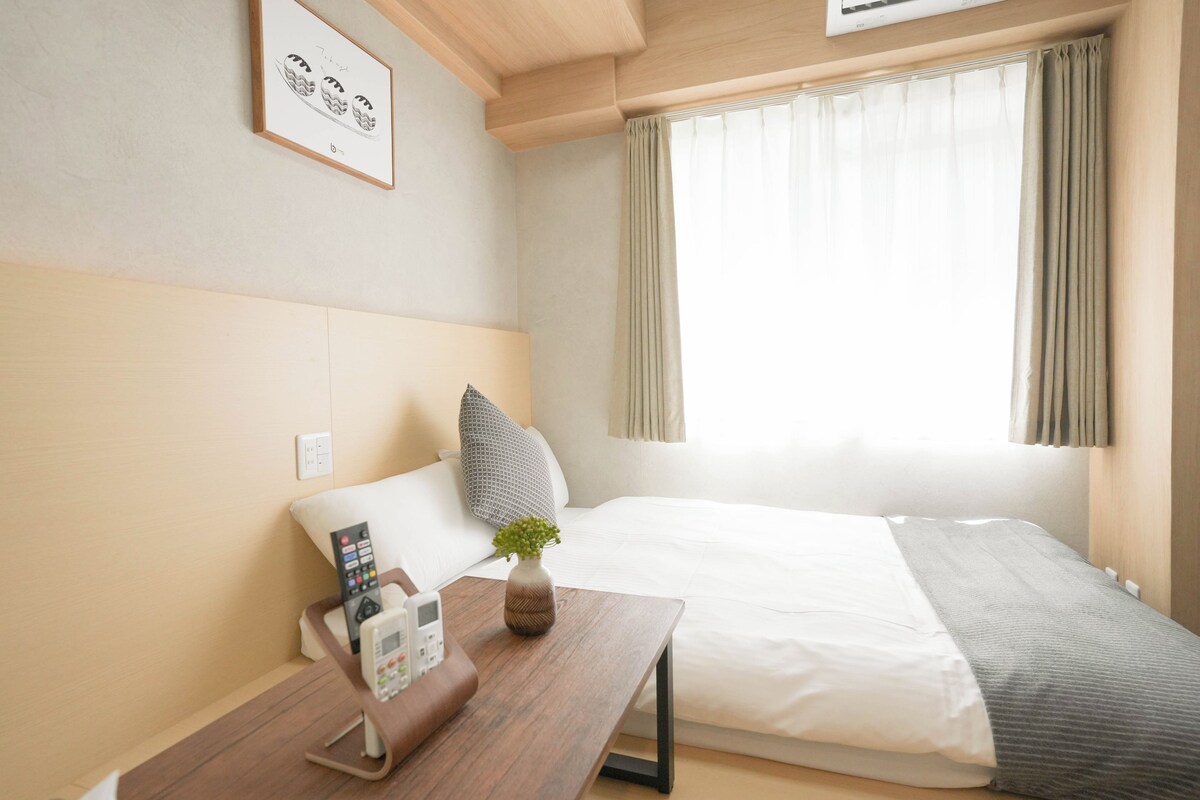 Ein Hotelzimmer mit einem Bett, einem Nachttisch mit Telefon, einer Pflanze in einer Vase und einem Fenster mit Vorhängen.
