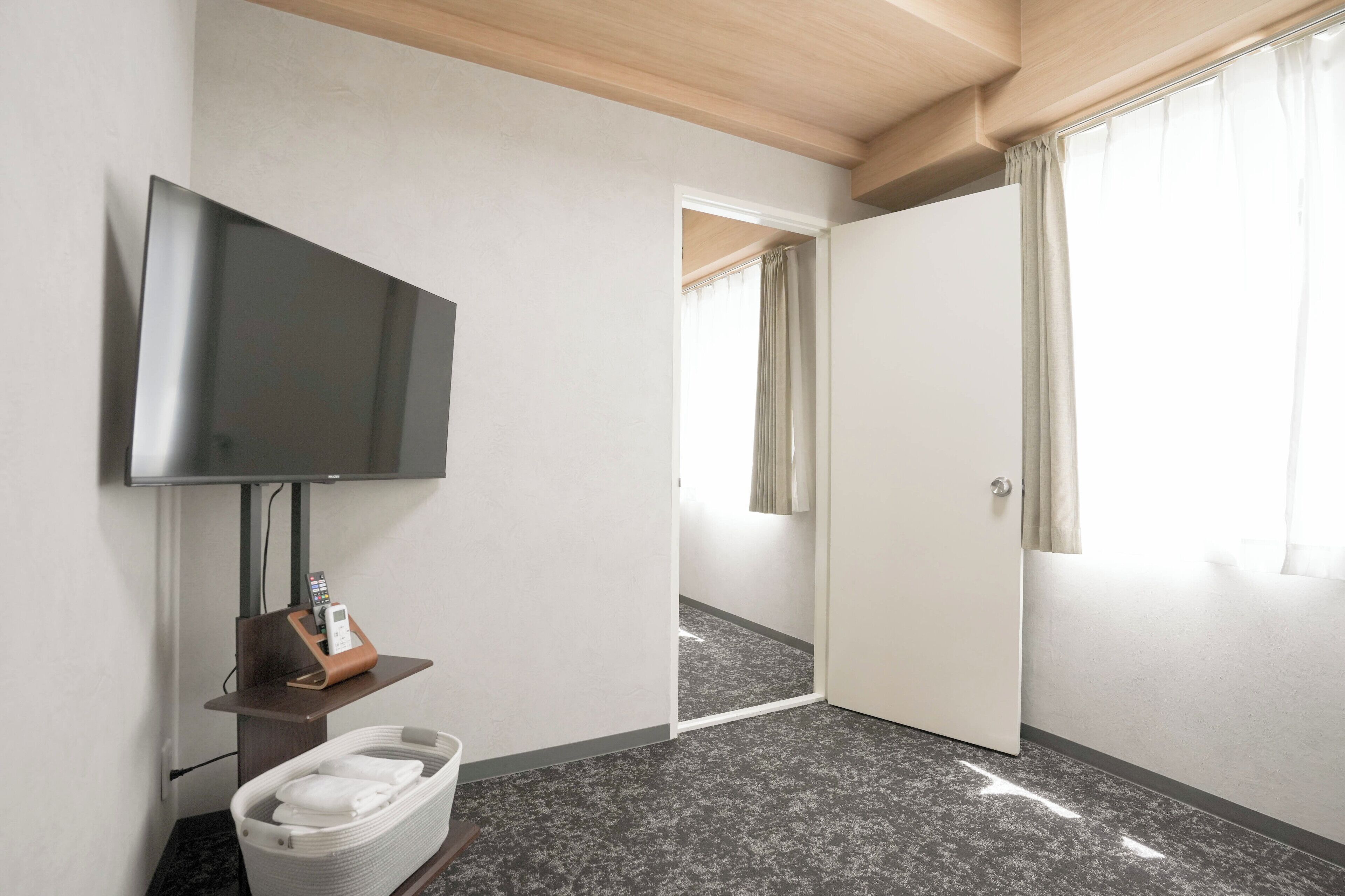 A hotel room with a flat-screen TV, a small table with a remote control, a basket with towels, and a door leading to another room.