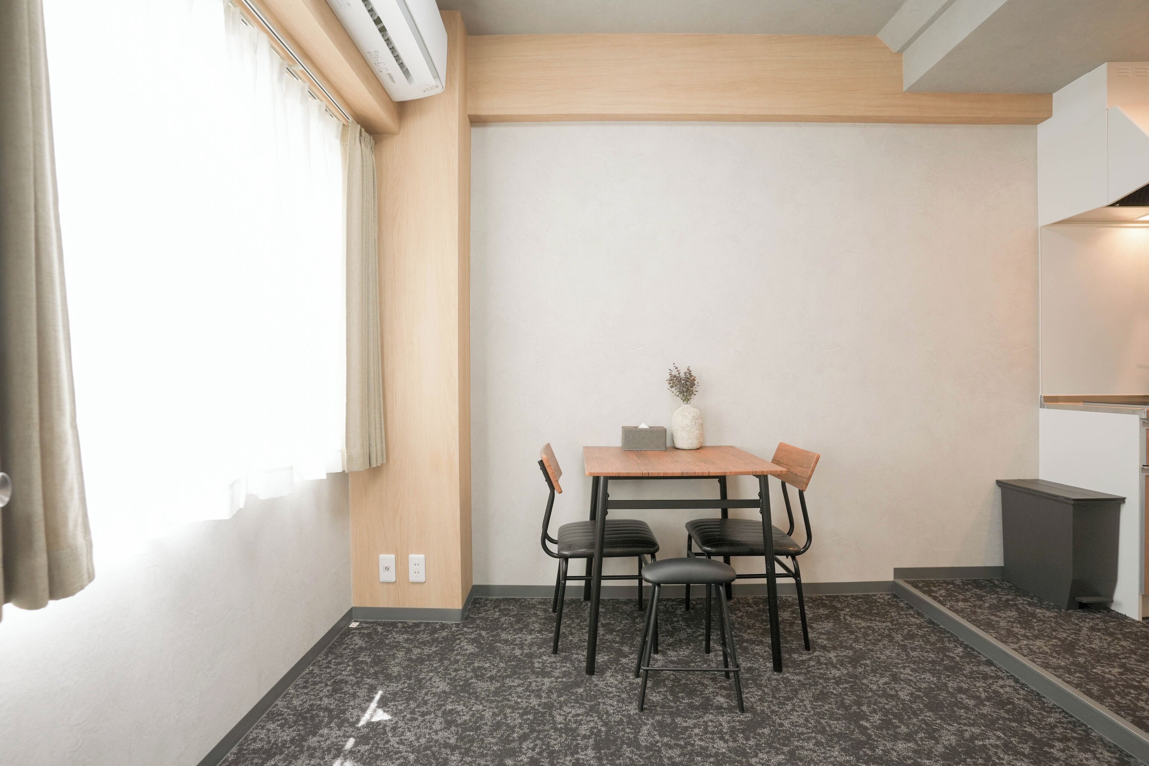 A small, modern room with a wooden table and four chairs, a small vase on the table, and a window with curtains.