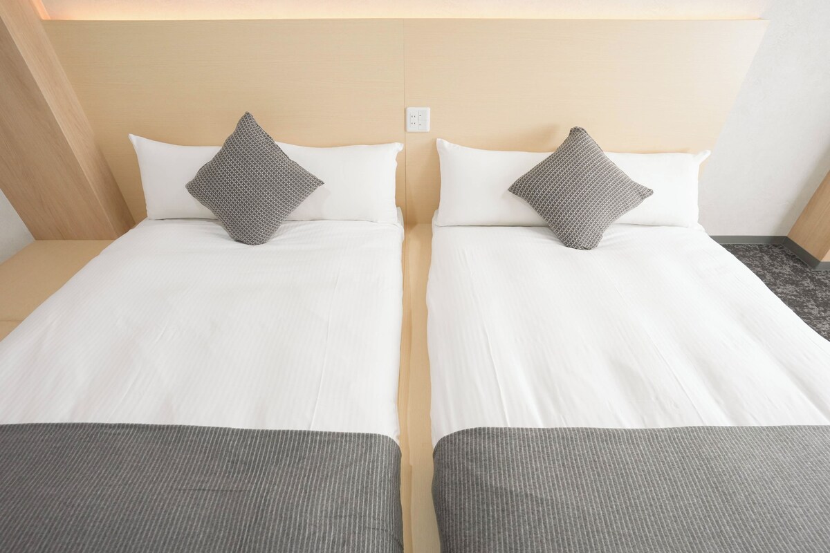 Ein Hotelzimmer mit zwei Einzelbetten, jeweils mit Kissen und einer grauen Decke.