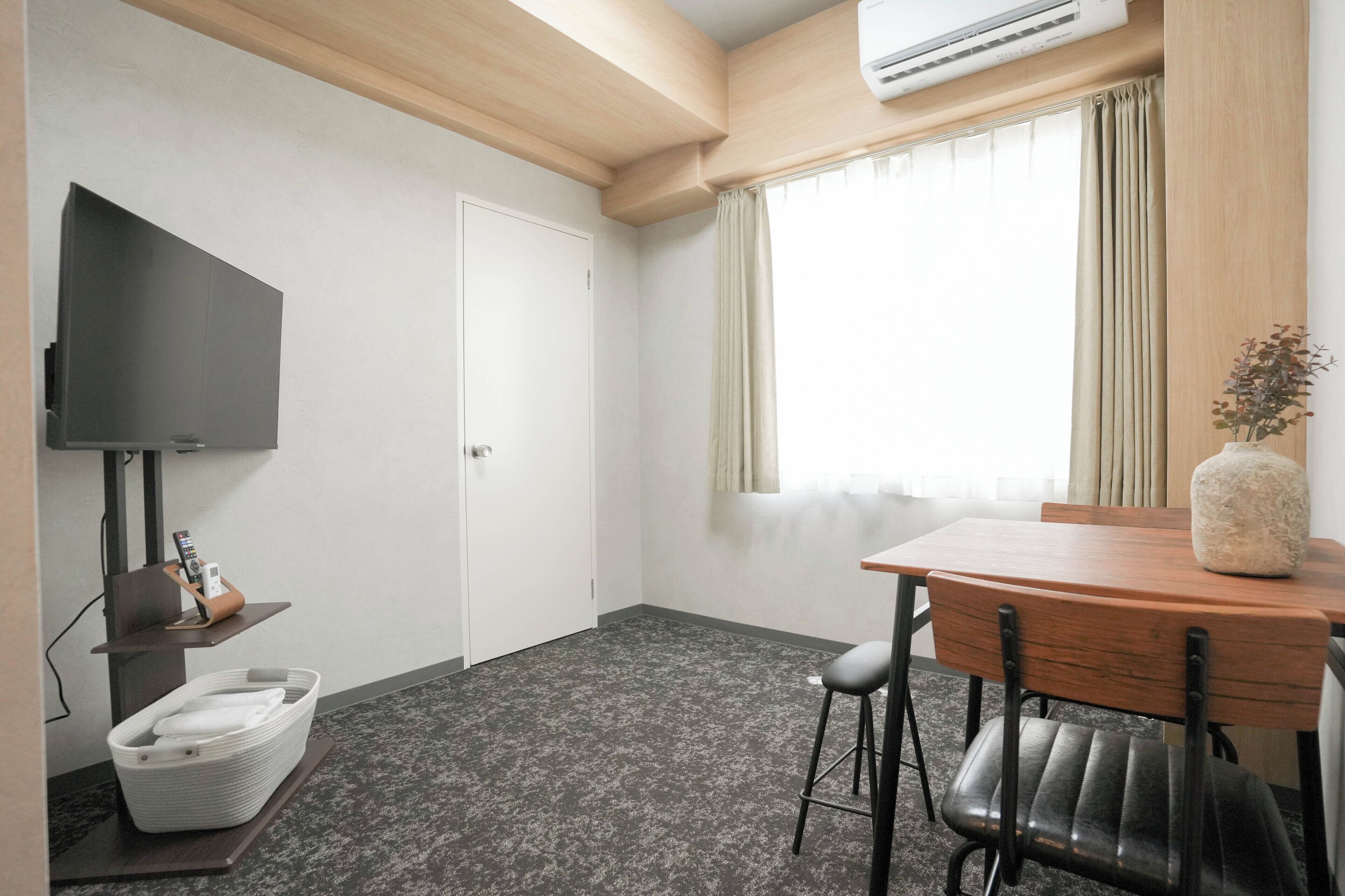 Habitación de hotel con televisor de pantalla plana montado en la pared, una mesa de madera con un florero, una silla, una cesta pequeña y una ventana con cortinas.