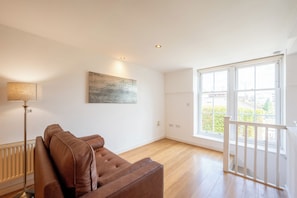 A spacious room with a brown leather sofa, a floor lamp, a painting on the wall, and a large window with a view of greenery. - Chic 1BDR Flat in De Beauvoir Town (London)