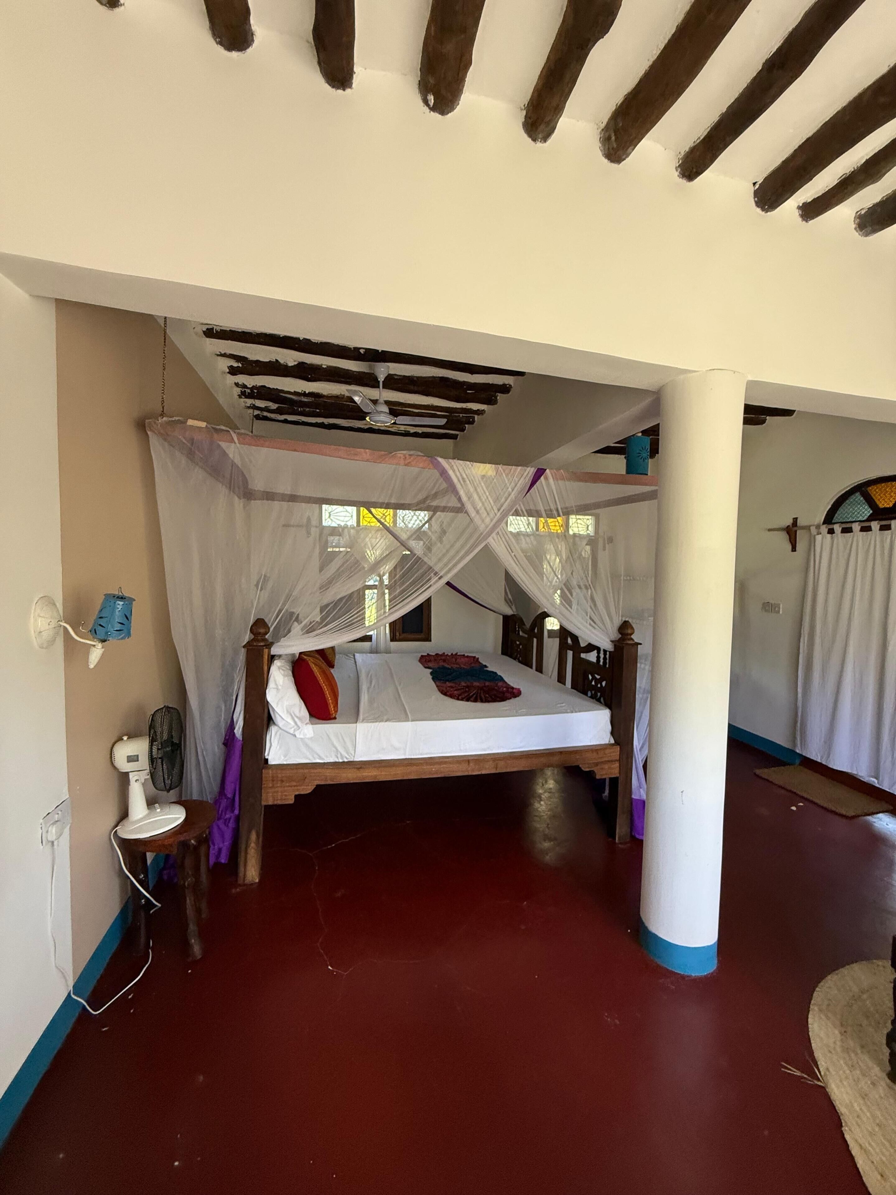 Une chambre avec un lit sous une moustiquaire, un ventilateur de plafond et un pilier en bois.