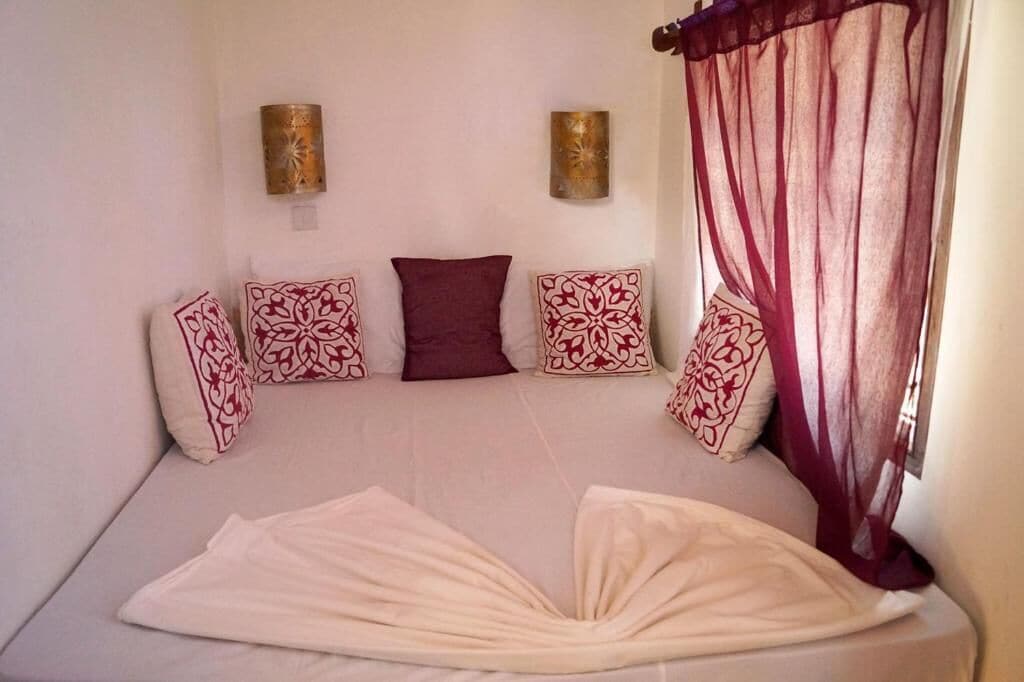 A room with a bed, four patterned pillows, two wall sconces, and sheer curtains.