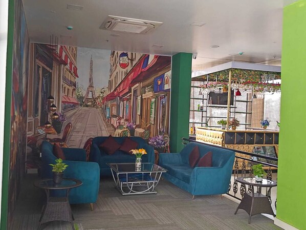 A room with a mural of a Parisian street, a blue sofa, a glass coffee table, and a small balcony with plants. - Inventree Hotel The Monarch Bagalur (Bengaluru)