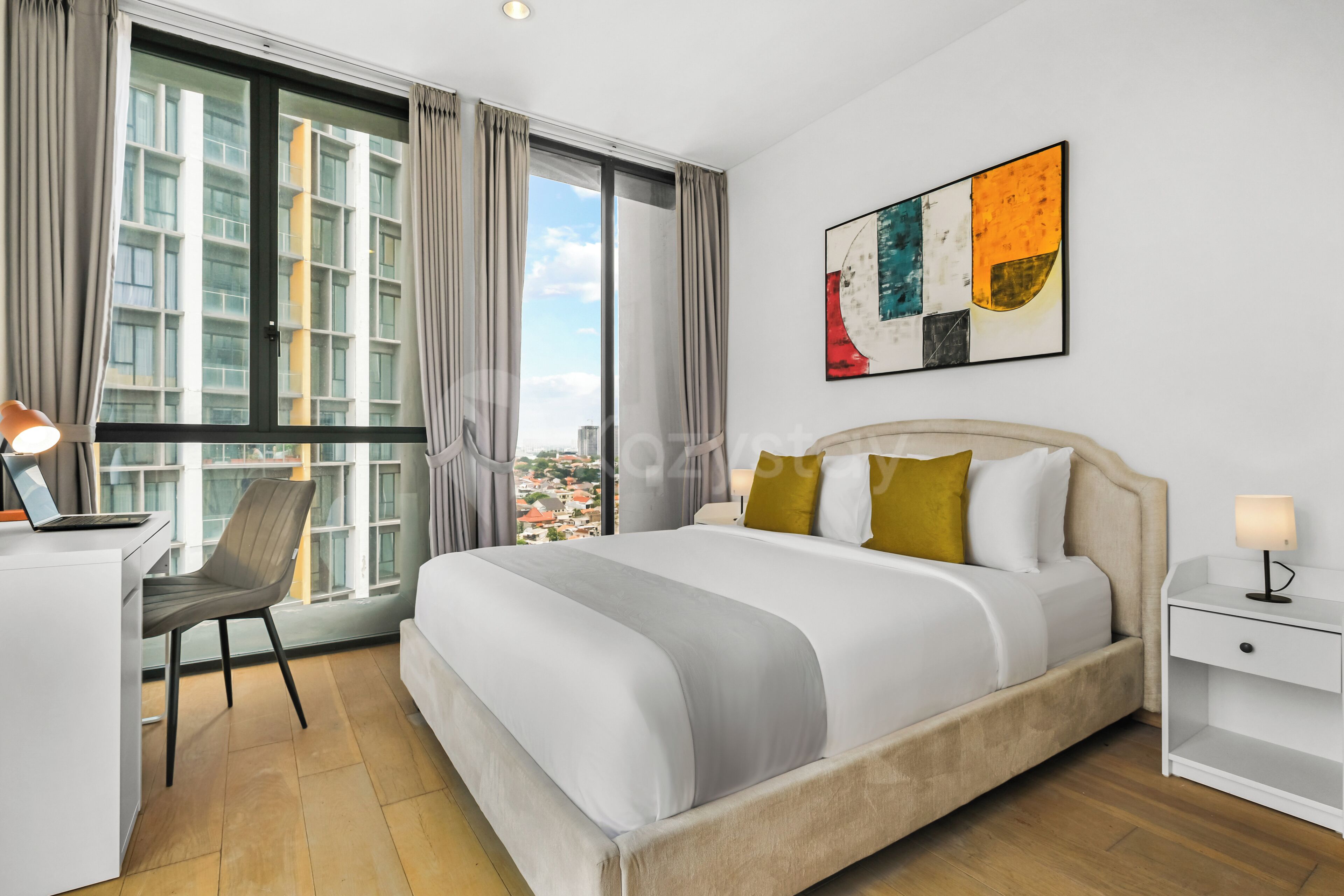 Habitación de hotel con cama, escritorio con computadora portátil, silla, mesita de noche con lámpara y un amplio ventanal con cortinas.