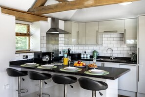 A modern kitchen with a central island, bar stools, and a window with blinds. - The Old Piggery at Rusty Lane Farm (Seend)