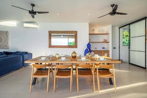 Ein moderner Essbereich mit einem Holztisch, gedeckt für sechs Personen, mit zwei Deckenventilatoren, einem blauen Sofa und einem großen Fenster mit Blick auf Grünflächen.