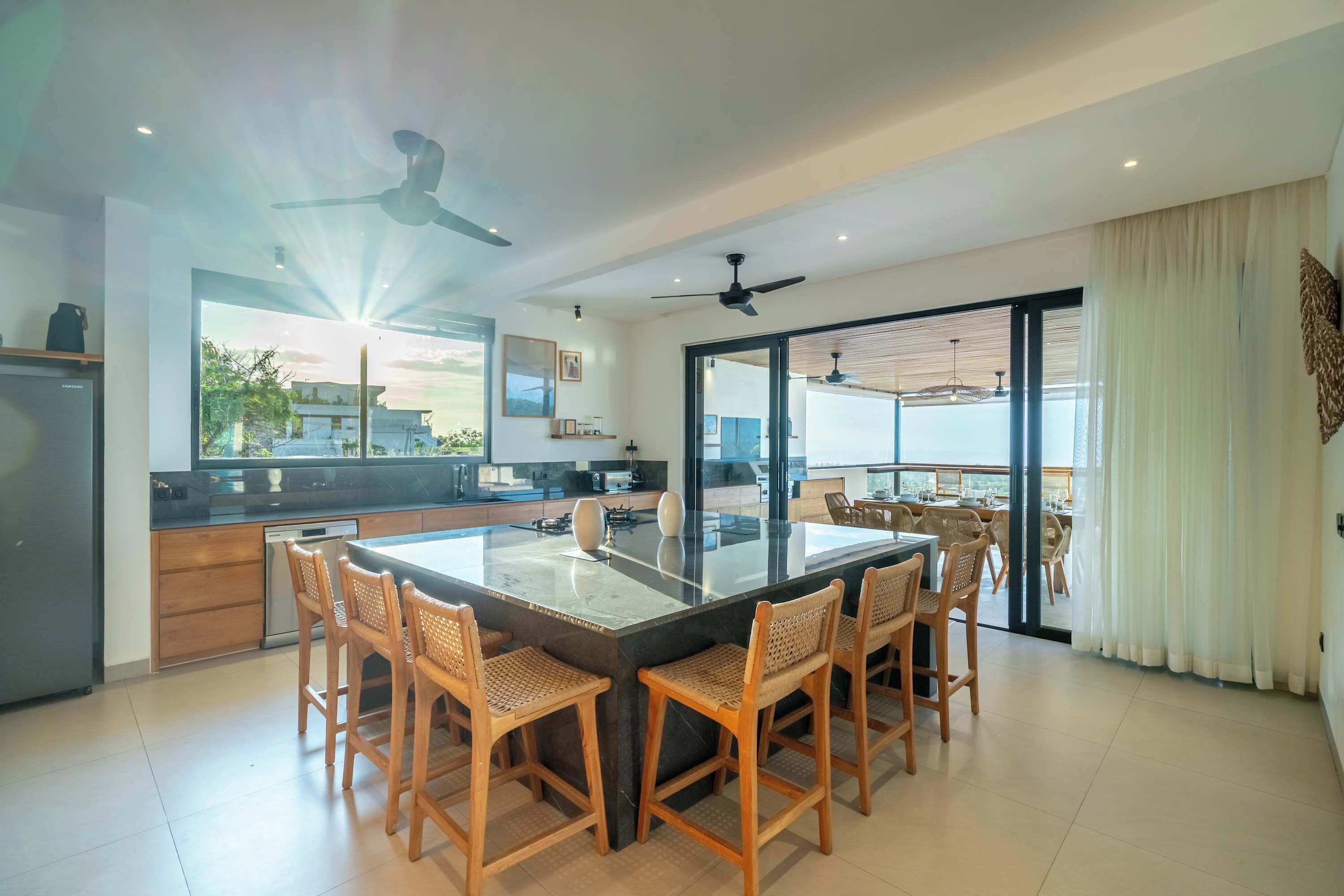 A modern kitchen with a central island, dining area with wooden chairs, ceiling fans, and large windows.