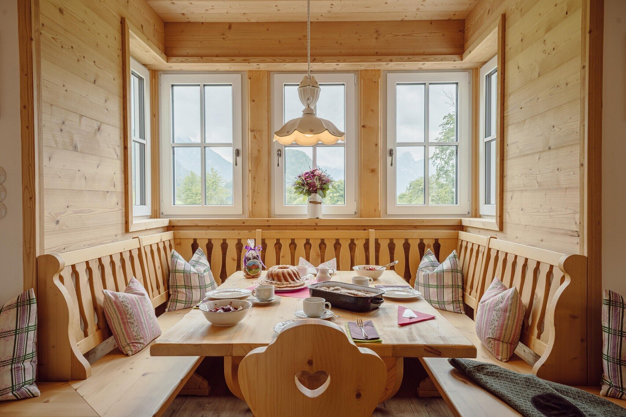 Et træbord med bænke, en hængende lampe og et vindue med udsigt til bjerge.