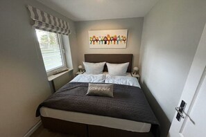 Ein Schlafzimmer mit Bett, einem Fenster mit gestreiftem Rollo, einem Wandbild und einem Nachttisch mit Lampe.