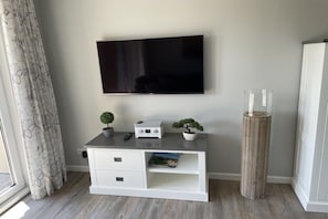 Uma sala de estar moderna com uma televisão de tela plana na parede, um móvel de entretenimento branco com gavetas, uma pequena planta em vaso, um vaso de madeira alto e um vaso de vidro no chão.