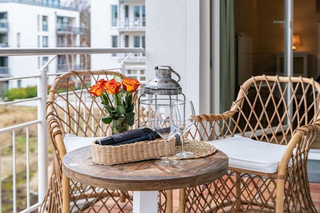 Una sedia e un tavolino in vimini con una bottiglia, bicchieri e fiori su un balcone.
