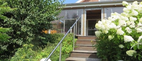 A house with a wooden staircase leading to a door, surrounded by lush greenery and white flowers.