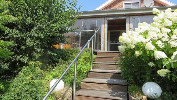 Ett hus med en trätrappa som leder upp till en dörr, omgivet av grönska och vita blommor.