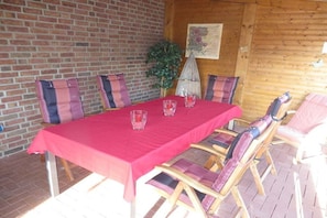 Una mesa de comedor de madera con mantel rojo, rodeada de sillas con cojines a rayas, sobre un patio con pared de ladrillo.