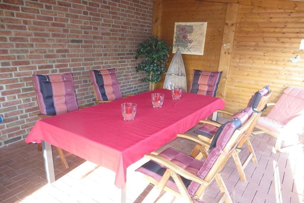 Una mesa de comedor de madera con mantel rojo, rodeada de sillas con cojines a rayas, sobre un patio con pared de ladrillo.