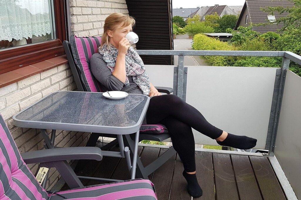 Une personne assise sur une chaise, devant une table, avec une fenêtre ornée de rideaux en dentelle, et une vue sur des maisons et de la verdure.