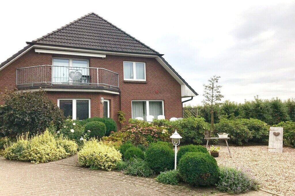 Une maison à deux étages dotée d’un balcon, entourée d’un jardin bien entretenu avec divers arbustes et une allée en gravier.