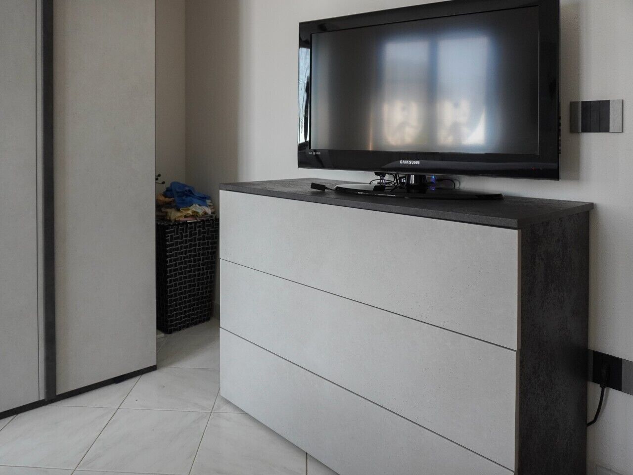 Una habitación moderna con una televisión de pantalla plana en la pared, un mueble gris con cajones y un cubo de basura negro con objetos.