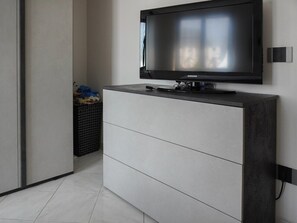 A modern room with a flat-screen TV mounted on a wall, a grey cabinet with drawers, and a black trash bin with items. - The Window Overlooking the Sea (Luni Mare)