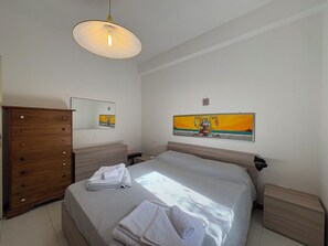 Une chambre à coucher comprenant un lit, une commode en bois, une table de chevet, une lampe de chevet, un tableau accroché au mur et un luminaire suspendu au plafond.