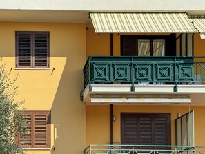 Ein Gebäude mit gelbem Anstrich, zwei Fenster mit Fensterläden, ein Balkon mit grüner Reling und ein gestreifter Markise.