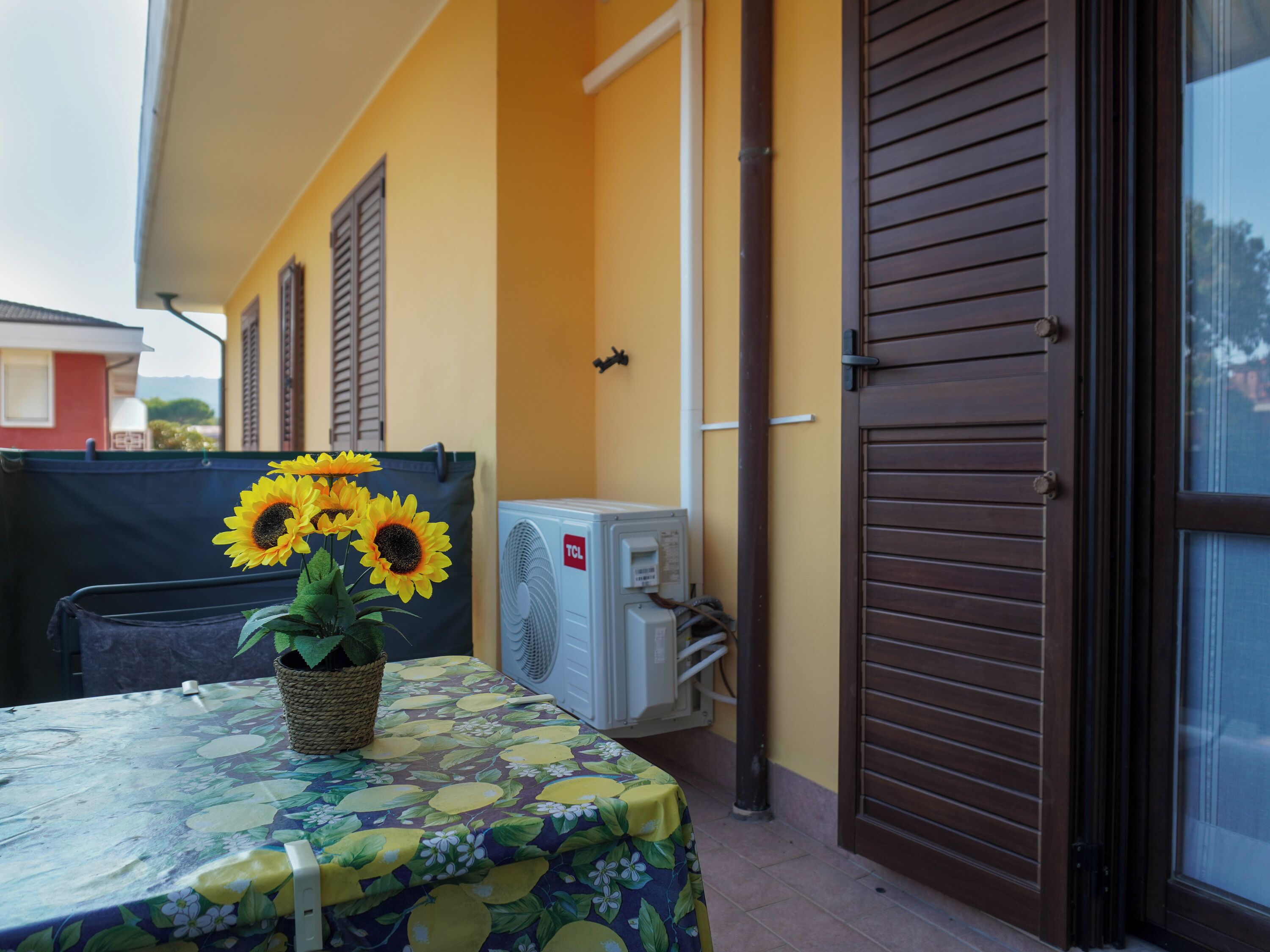 Ein Balkon mit Tisch, bedeckt mit einer blumigen Tischdecke, einer Topfpflanze mit Sonnenblumen und einer Klimaanlage.