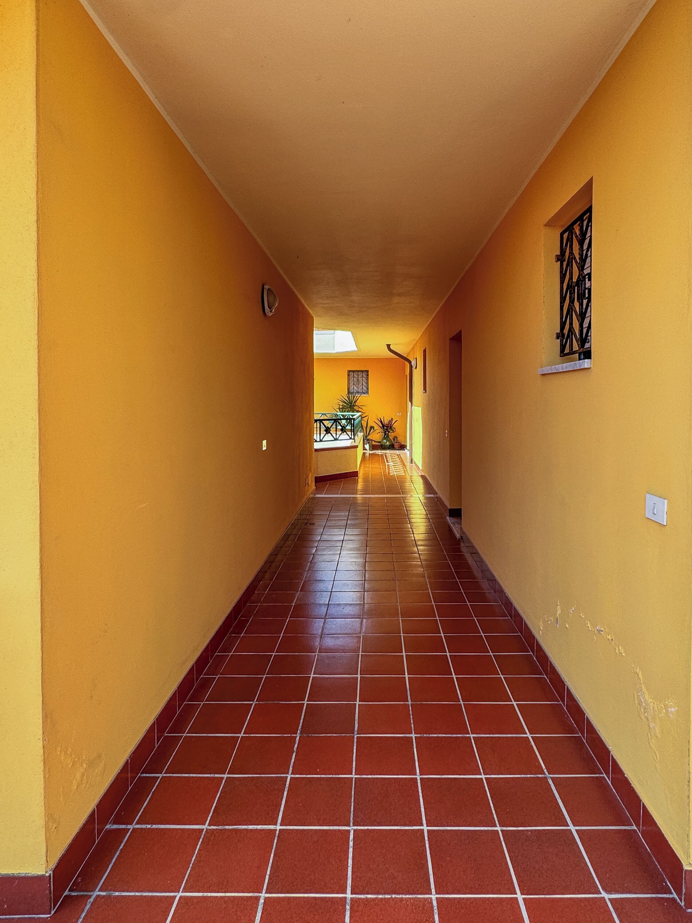 A corridor with yellow walls and red-tiled flooring, leading to a room with a window and a plant.