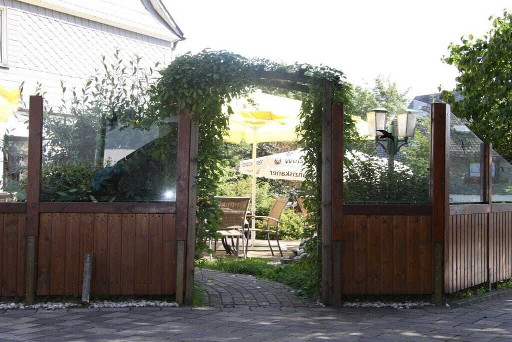 Una verja de madera con un porche cubierto de vegetación, que da acceso a una zona de estar con sillas y una mesa.