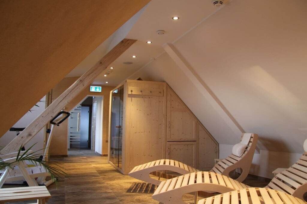 A room with a sloped ceiling, wooden flooring, and a staircase leading to an upper level. There are wooden chairs and a bench along the wall.