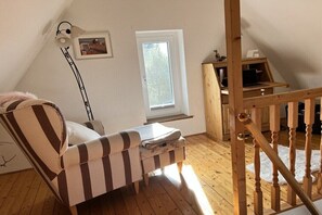 A room with a sloped ceiling, a wooden desk with a laptop, a striped armchair, a floor lamp, and a window. - Holiday home in Dranske with swimming pool (Dranske)