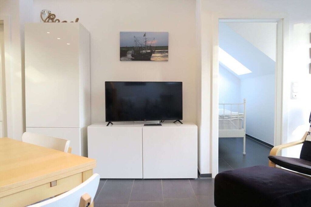 A modern living room with a flat-screen TV on a white cabinet, a dining table with chairs, and a view of a balcony with a railing.